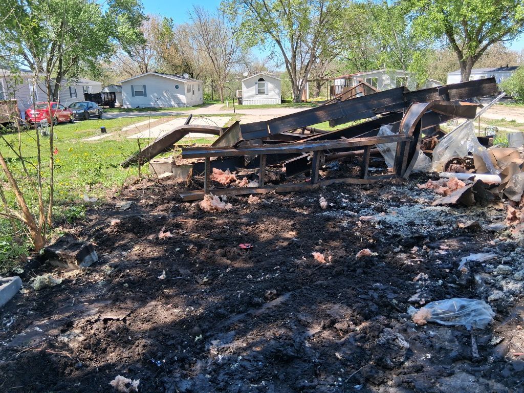 Burned debris and charred remains of a collapsed structure in a yard, with trees and houses in the background