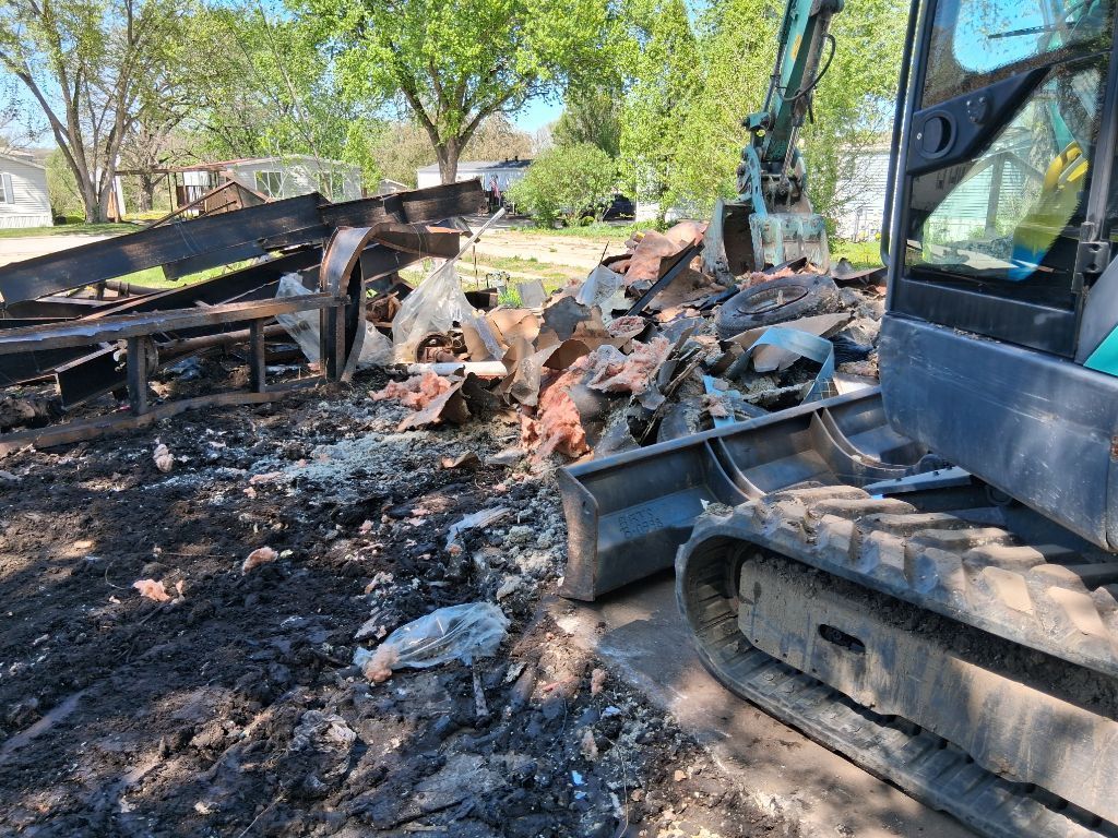 Burned debris and a damaged excavator at a fire-damaged site with charred ground and rubble