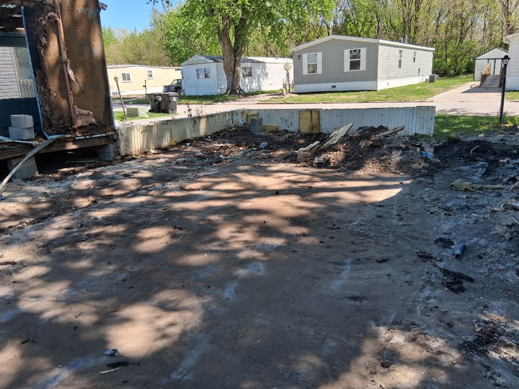 Burned ground and debris beside a large tree, with mobile homes in the background.
