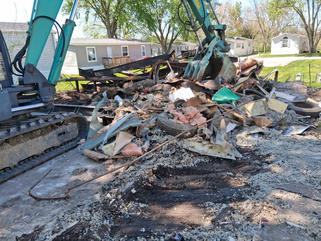 Excavator clearing a burned debris pile in a suburban yard, with smoke and charred remains visible.