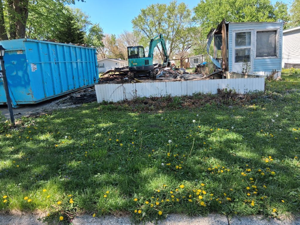 Blue dumpster beside a white trailer and small excavator on a grassy lot with trees and yellow flowers