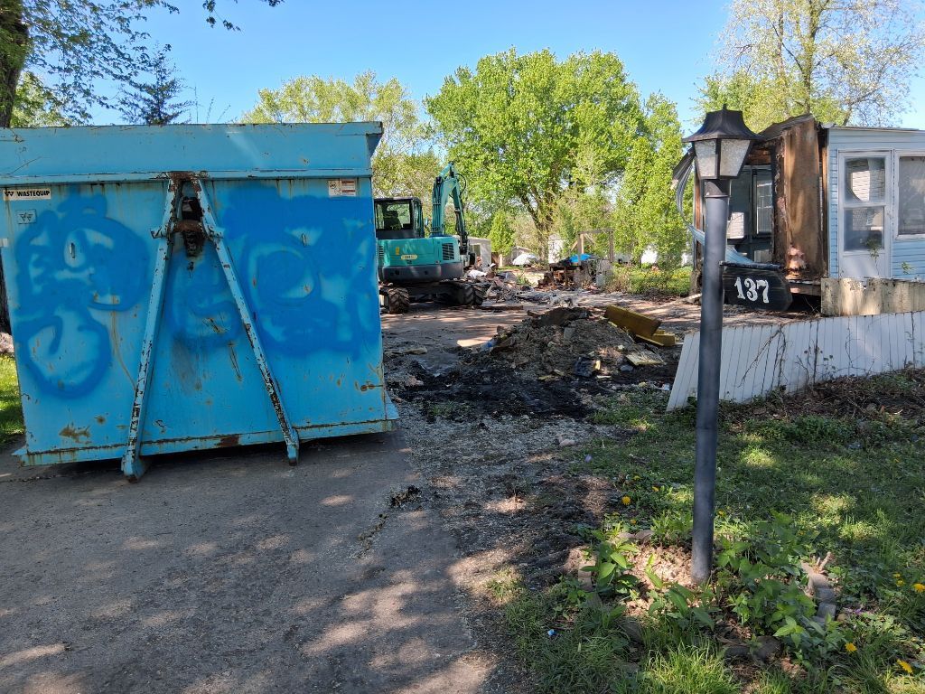 Blue dumpster in a yard with a parked truck and debris near a house gate.