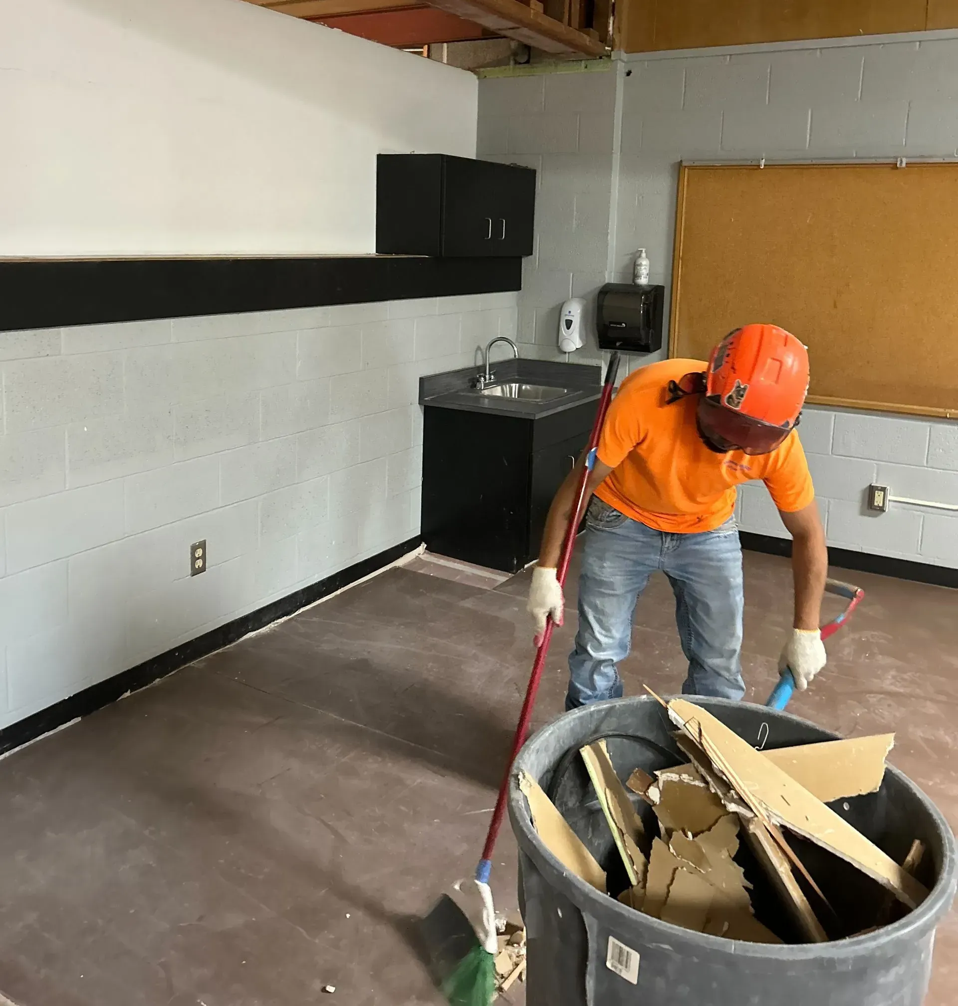 A person in an orange shirt and hard hat sweeps construction debris into a large plastic bin inside a room.