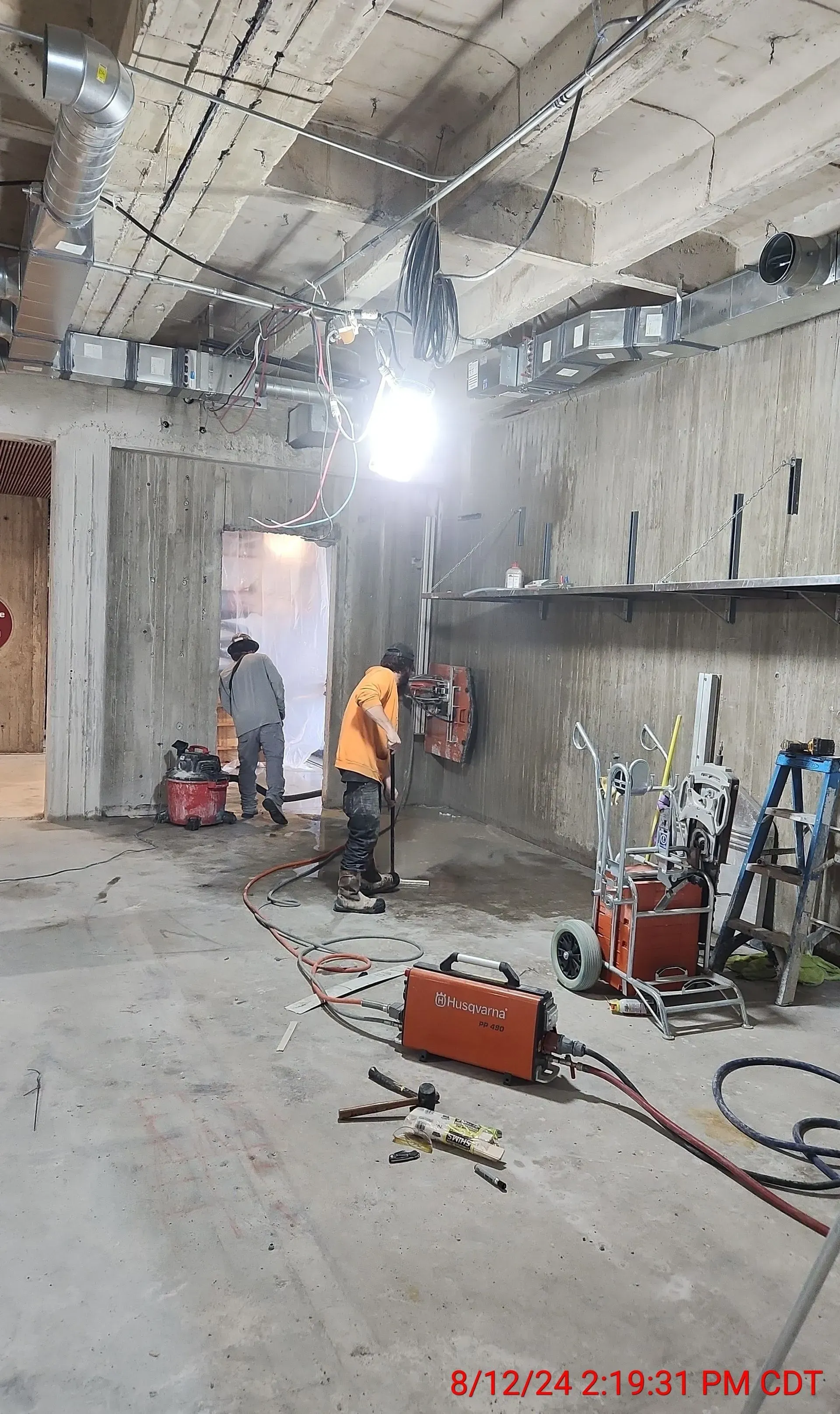 Two workers in a construction site operate concrete cutting equipment near a wall, with tools and a bright light nearby.