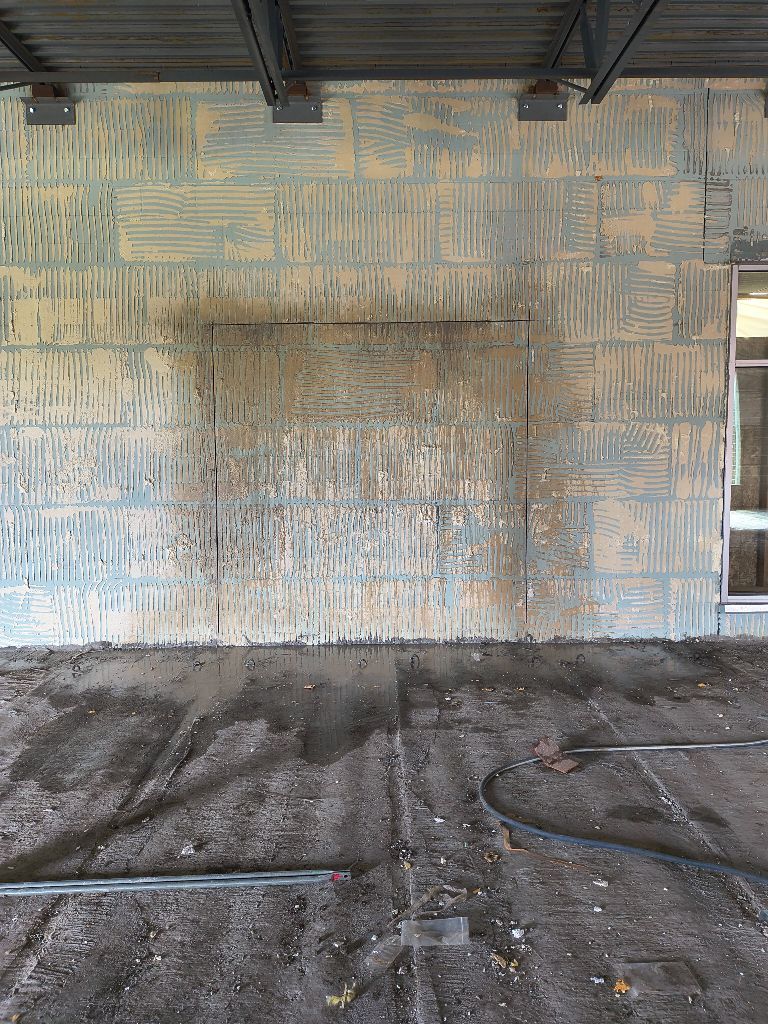 Weathered concrete wall under a roof, with a stained floor and dark hose or cable on the ground.