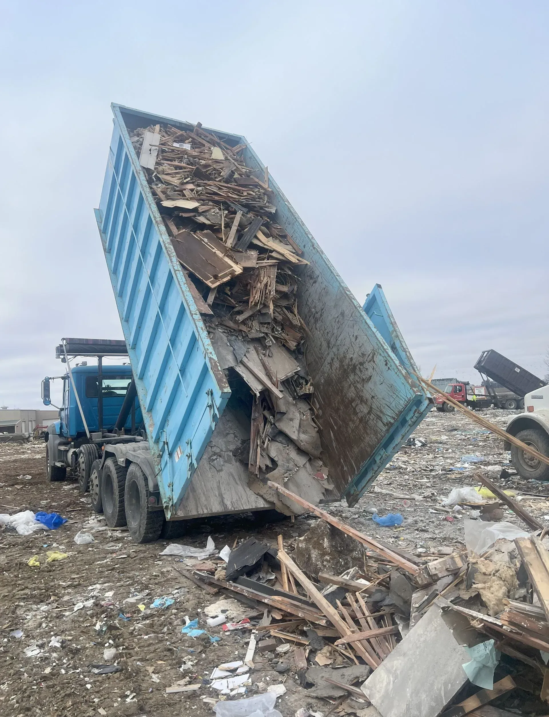 A blue dump truck in a landfill, tilted at a steep angle to discharge a load of construction debris onto the ground.