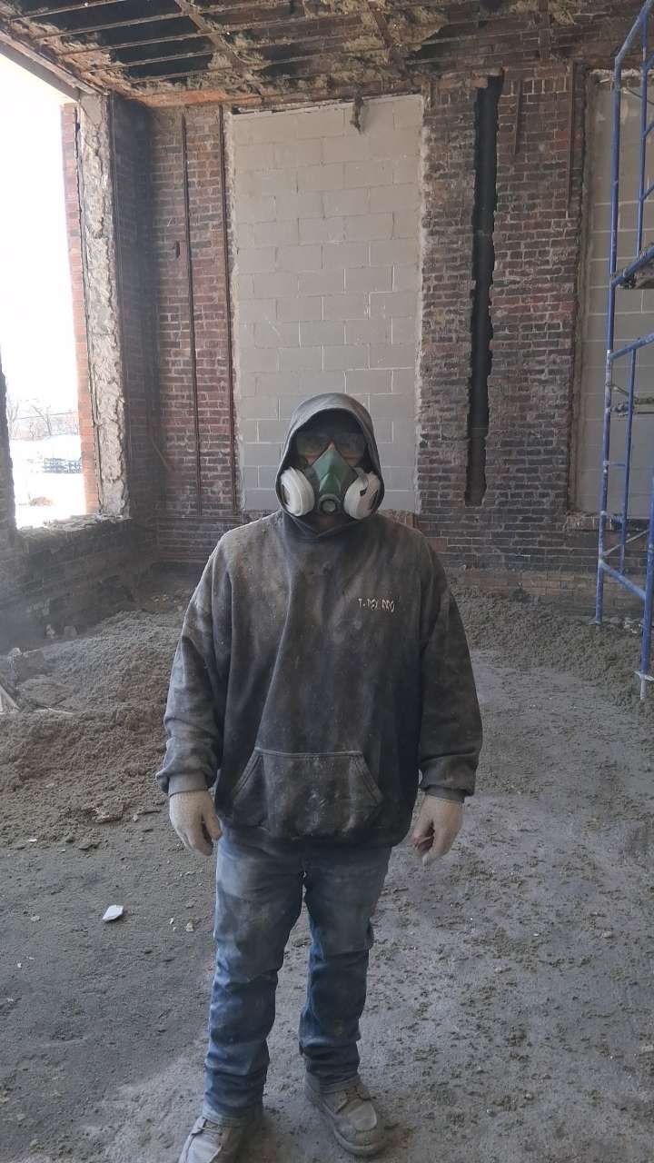 Person in a gas mask standing in an unfinished brick building with a bare concrete floor.