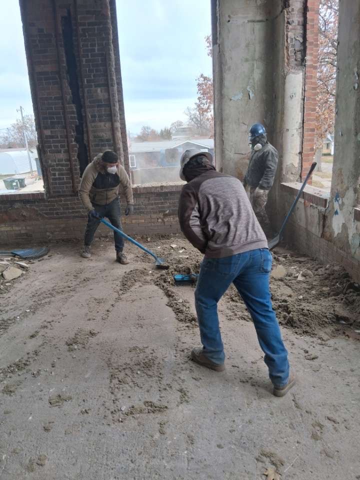Three workers shoveling debris inside an unfinished brick building with large window openings