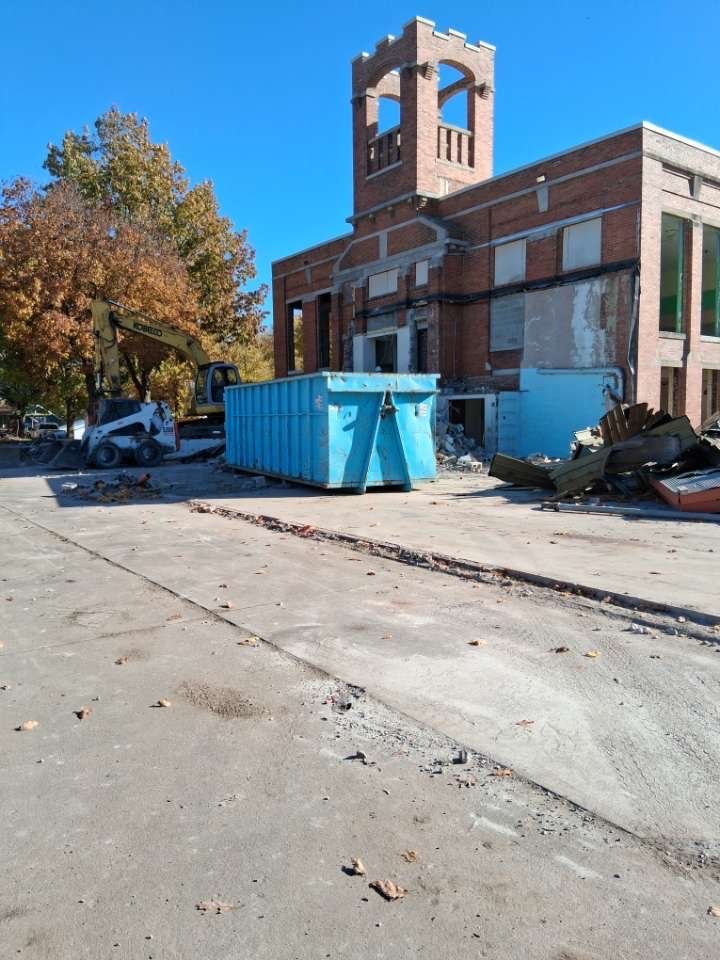 Blue dumpster beside a brick building and bell tower on a sunny street, with parked cars and scattered debris