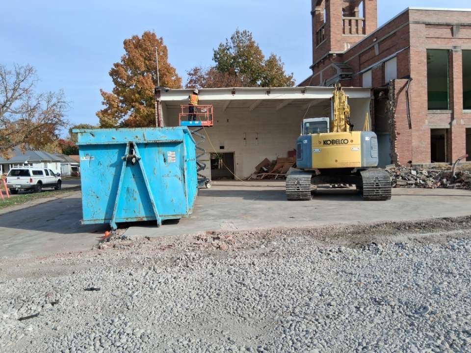 Construction site with blue dumpster and yellow excavator beside a brick building