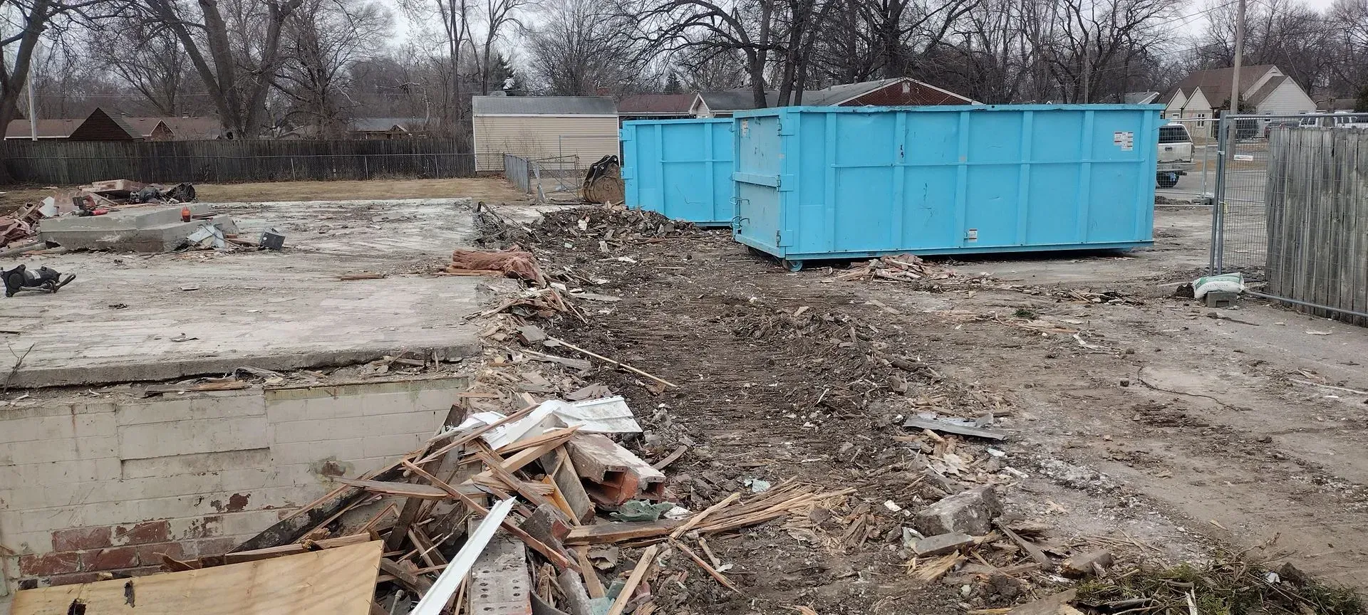 Two bright blue dumpsters sit at a construction site filled with concrete rubble and scattered building debris.