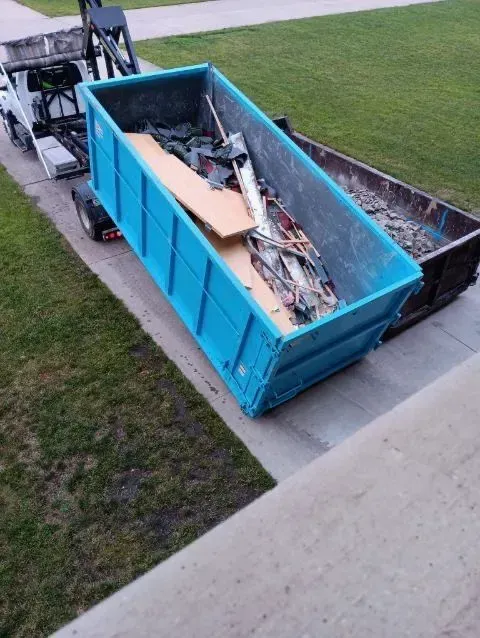 A bright blue dumpster filled with construction debris sits on a concrete driveway, being loaded onto a truck.