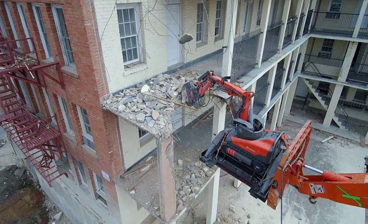 An orange demolition machine tears down a section of a multi-story brick building, leaving exposed rubble.