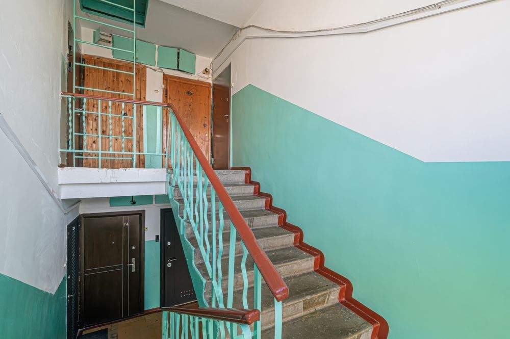 A stairwell with light green walls, a rusty brown handrail, grey stone steps, and two dark metal doors on the landing.