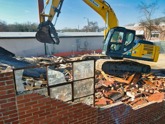 A teal excavator sits in front of a brick building wall undergoing partial demolition, revealing a concrete sub-structure.