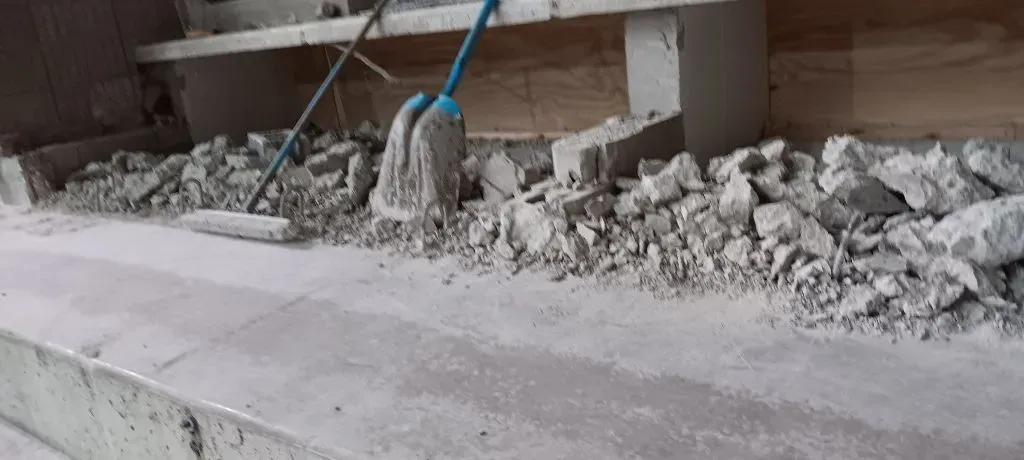 A blue-handled mop rests against a wall behind a pile of gray concrete rubble and debris on a concrete floor.