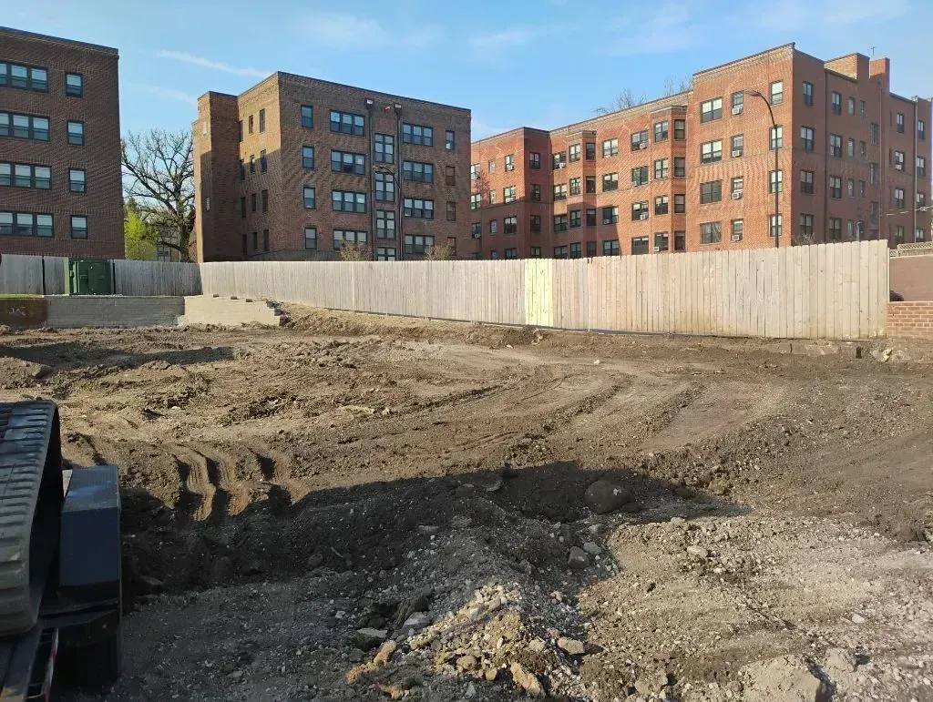 A cleared construction site with piles of dirt, bordered by a wooden fence in front of several brick apartment buildings.