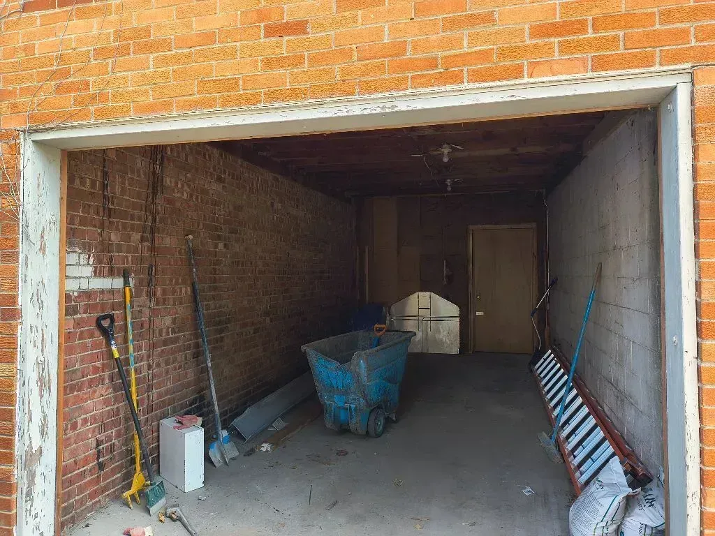 An open garage with exposed brick walls, containing a blue plastic bin, a ladder, and assorted tools on the concrete floor.