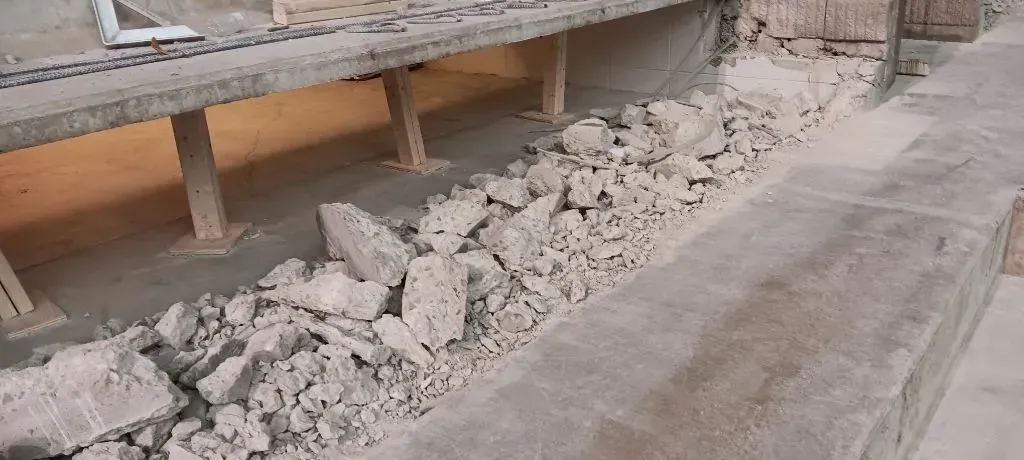 A trench filled with piles of grey concrete rubble, situated beneath a raised concrete walkway at a construction site.