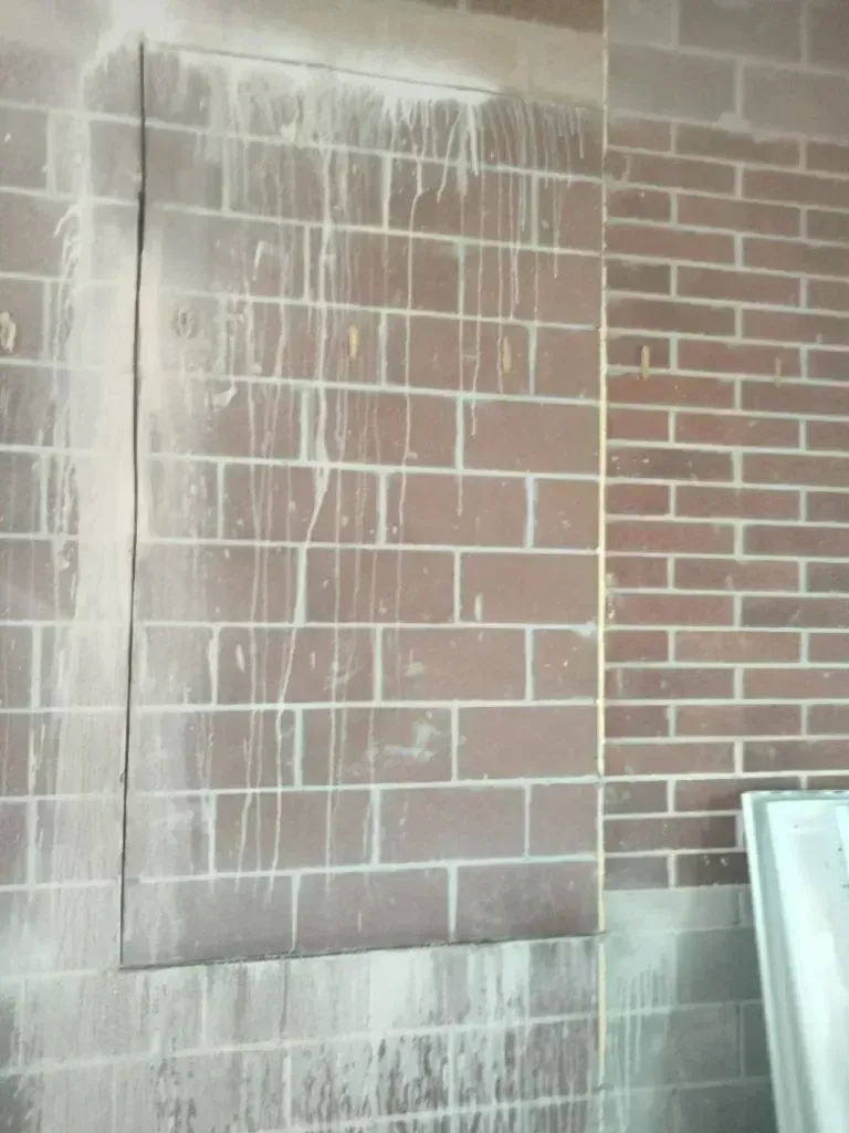 A brick wall with a rectangular metal access panel covered in streaks of dried white liquid residue.