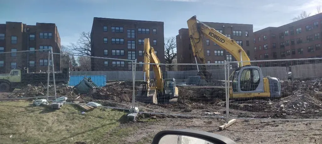 Two yellow excavators work on a dirt construction site in front of multi-story brick apartment buildings.