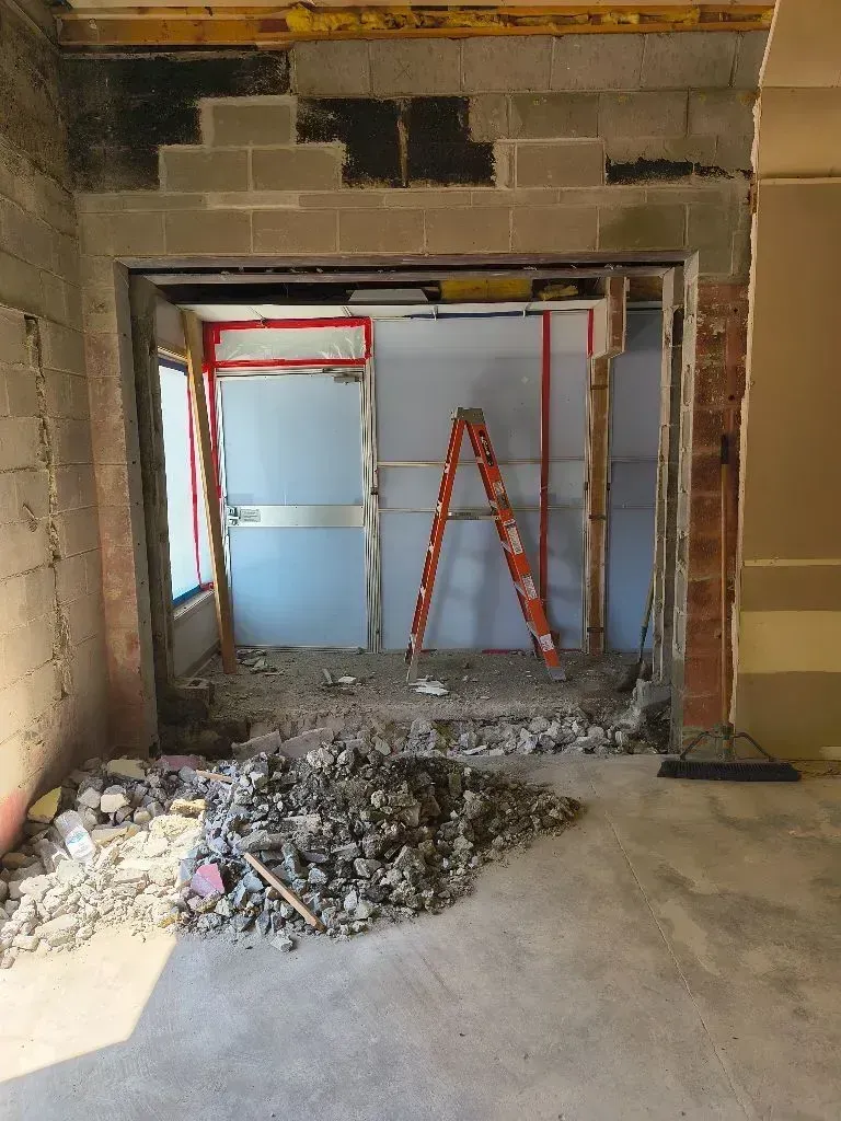 Construction area with a hole cut into a concrete block wall, rubble on the floor, and a ladder inside the opening.