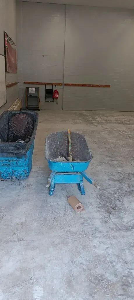 A blue wheelbarrow and a plastic container sit on a dusty concrete floor in an unfinished, empty room.