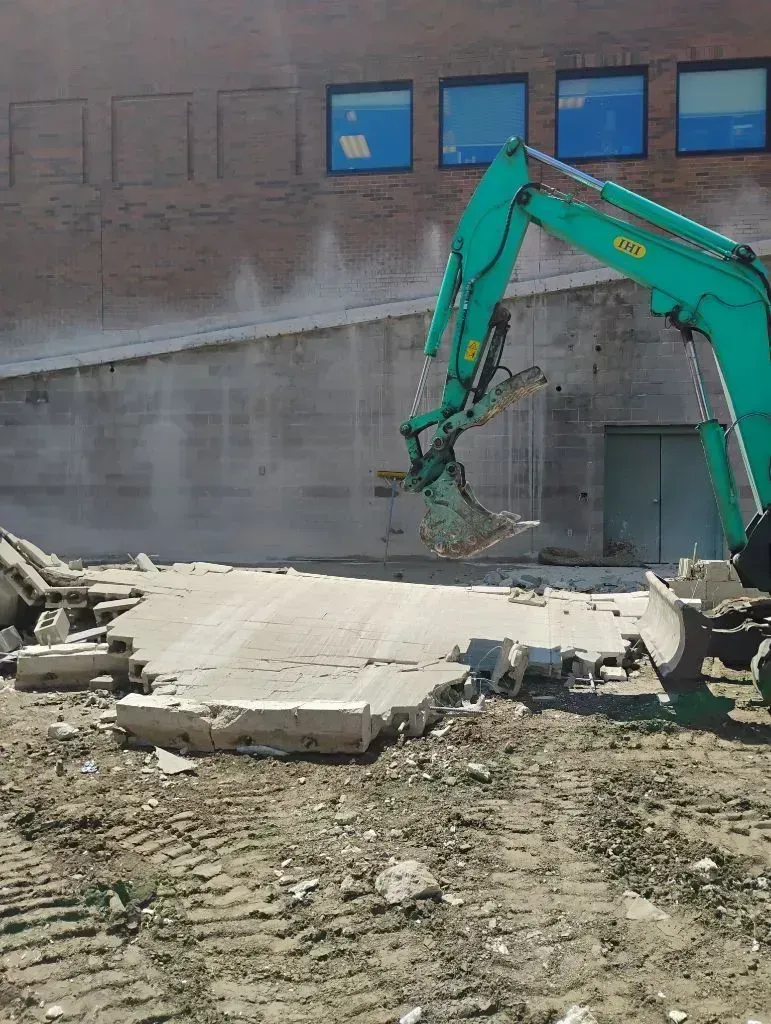 A teal construction excavator breaking up a concrete slab next to a brick building.