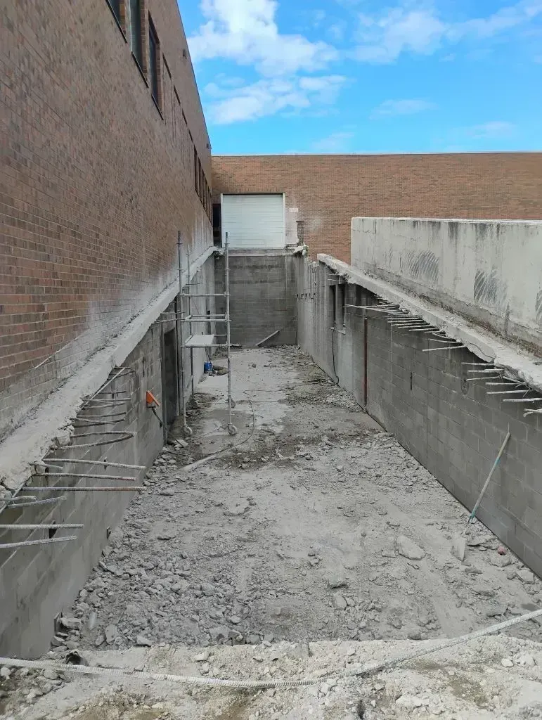 Construction site with a deep excavated trench between two brick walls and concrete footings with exposed rebar.