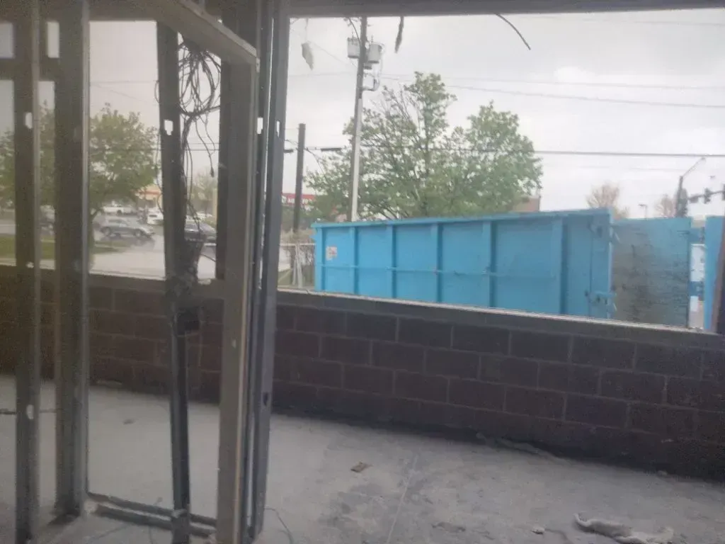 Interior view through a metal-framed doorway showing a brick wall and a large blue dumpster outside.