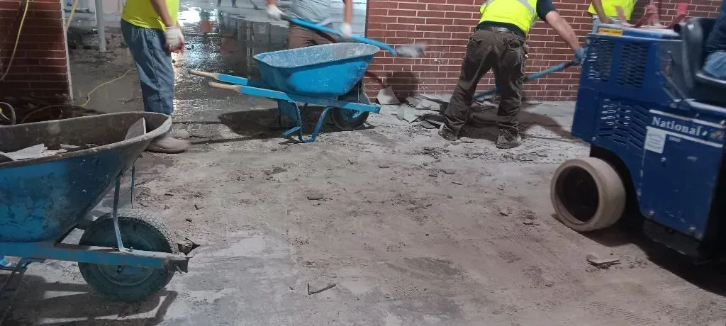 Construction workers use blue wheelbarrows and machinery to clear debris on a job site with a brick wall in the background.