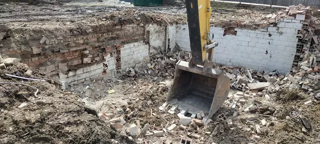 An excavator bucket sits amidst construction debris and exposed, painted brick walls at a demolition site.