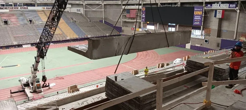 A crane lifts a large concrete structure inside an empty sports arena during construction.