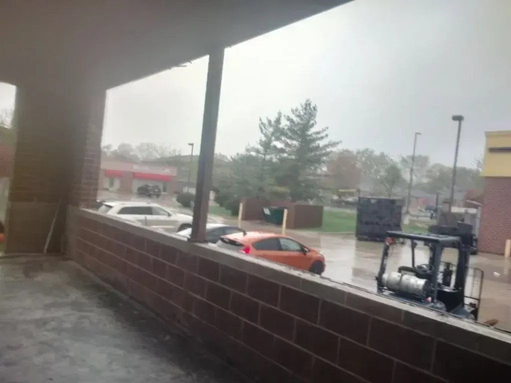 A rainy day view from under a porch looking at a parking lot with several cars, a forklift, and trees in the distance.