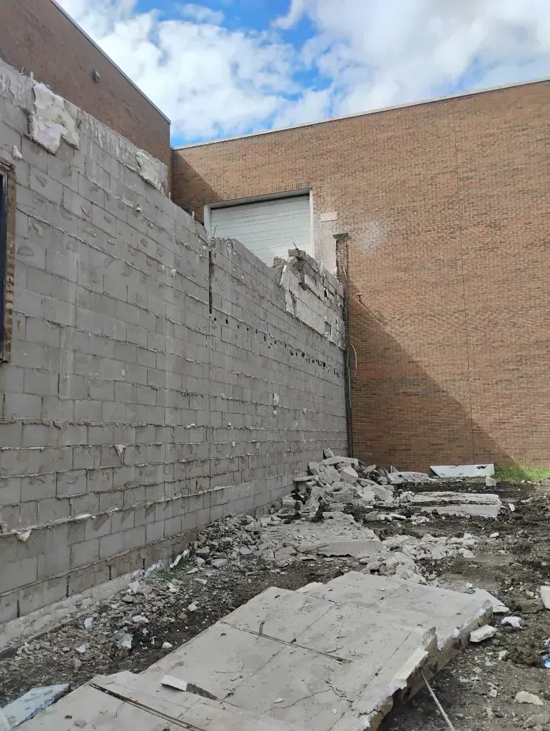 A partially demolished gray concrete block wall stands next to a tall, reddish-brown brick building under a cloudy sky.