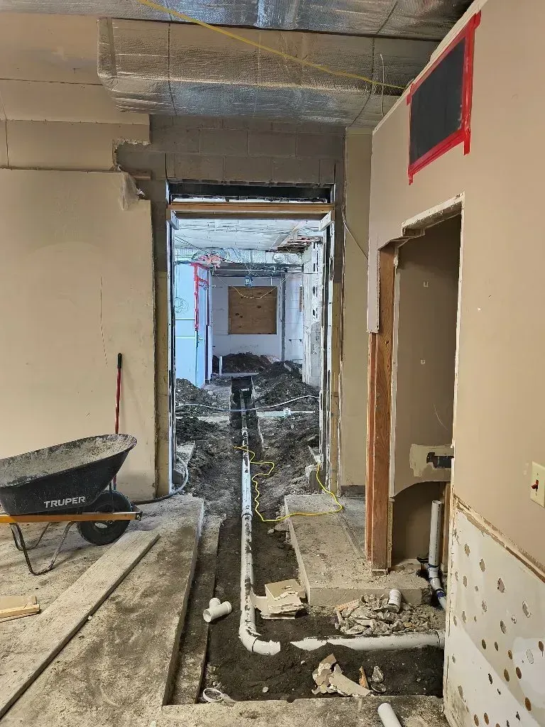An indoor construction site shows a trench cut into the concrete floor with exposed white plumbing pipes, near a cart.