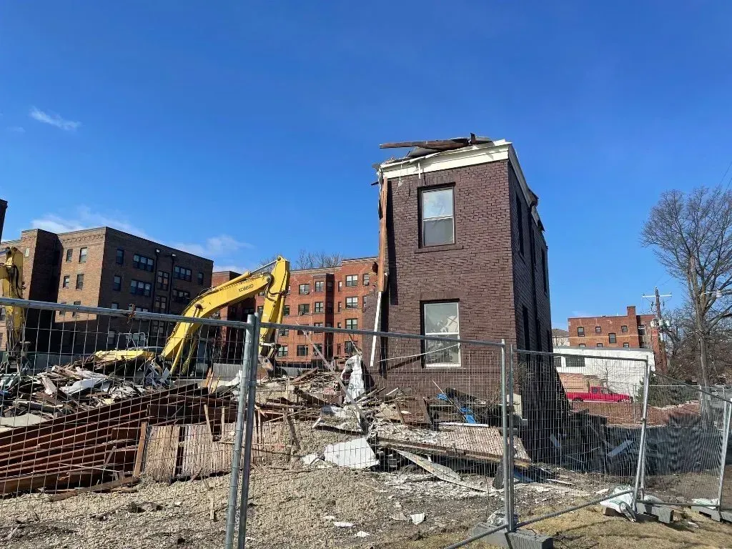 A yellow excavator demolishes a brick building, with a solitary wall left standing amidst debris under a clear blue sky.