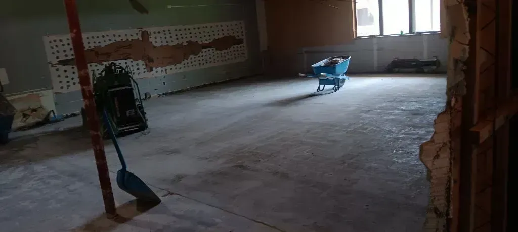 An interior view of a room under construction, showing a concrete floor, a blue wheelbarrow, and a blue shovel.