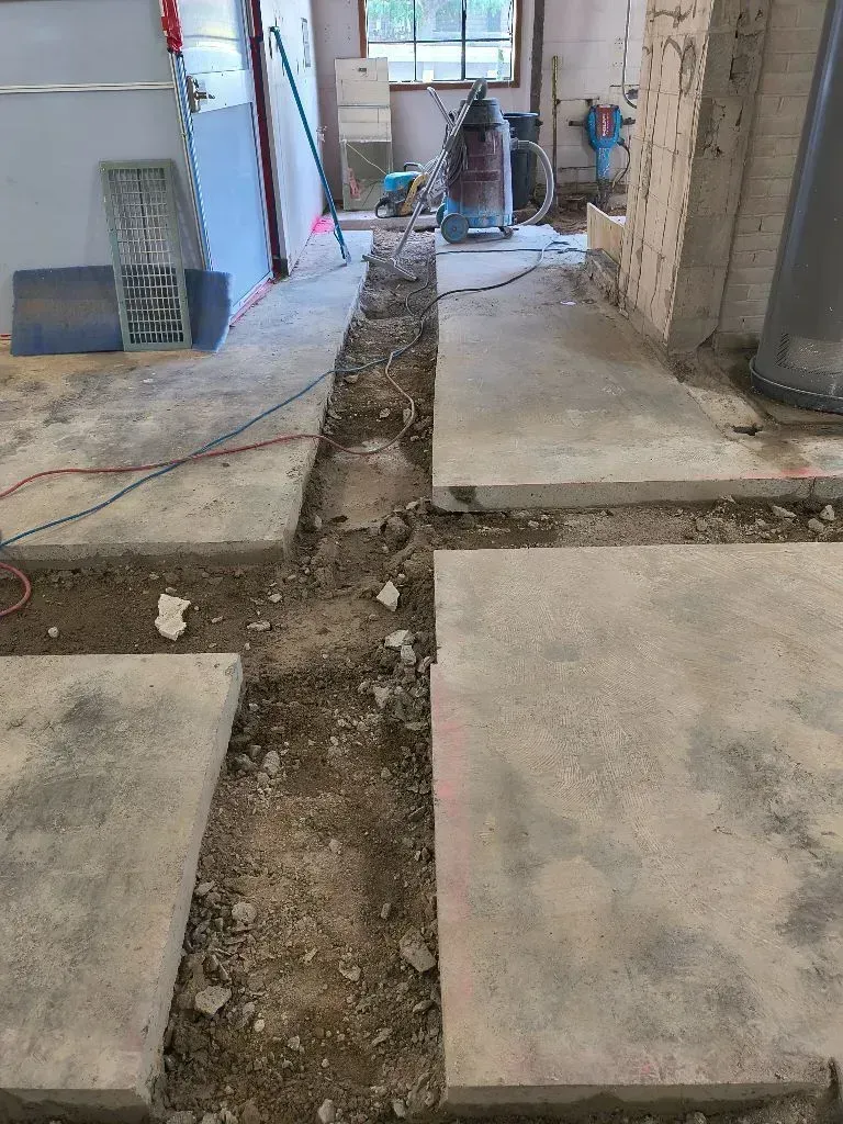 A trench cut through a concrete floor in a room under construction, with tools and debris visible in the background.