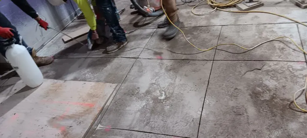 Construction workers stand on a concrete floor while spraying water on marked cuts in the surface.