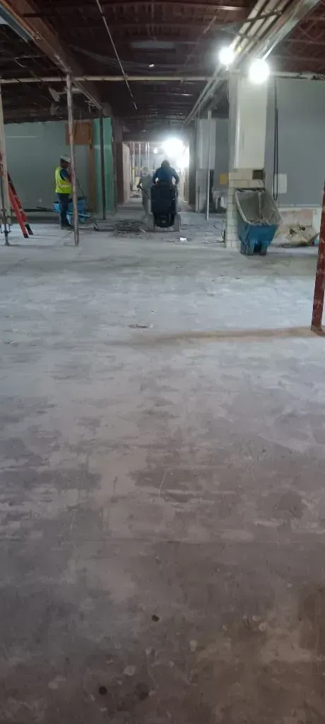 Construction workers inside a large, unfinished industrial space with concrete floors, exposed framing, and bright lights.