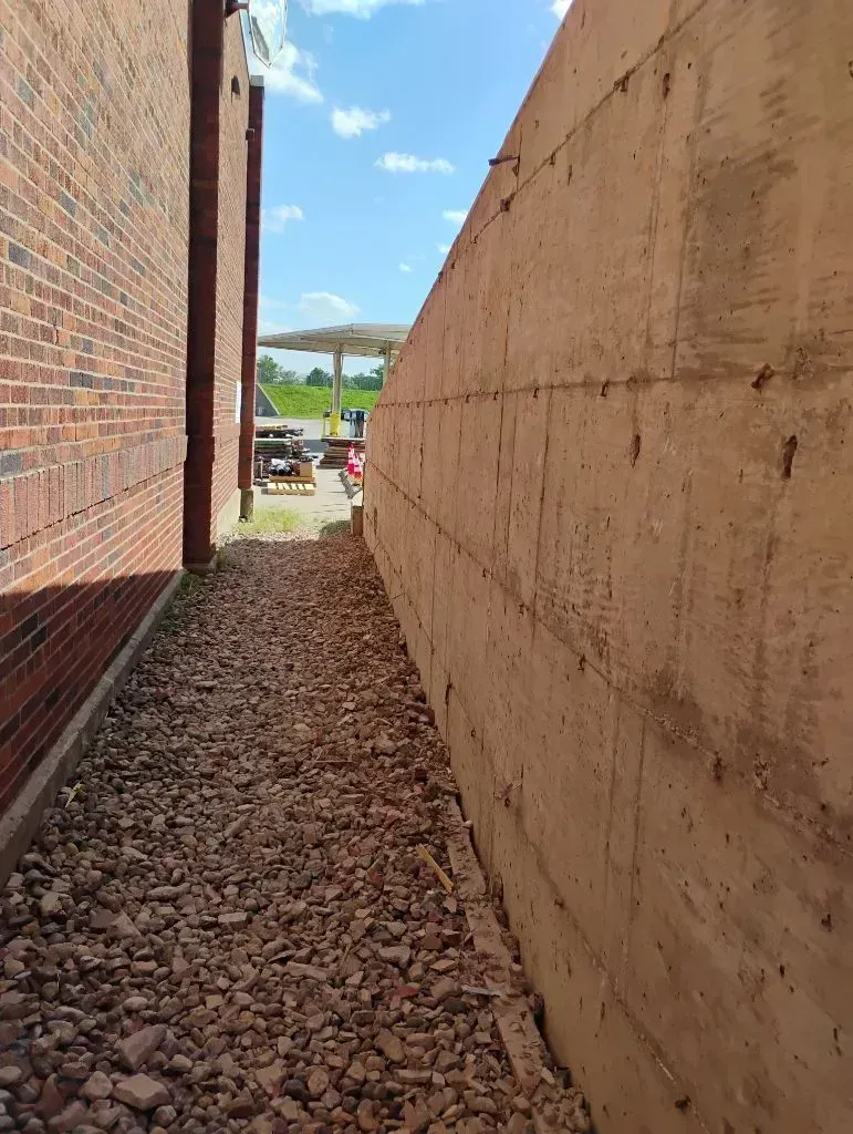 A narrow outdoor walkway with red brick on the left, a tall concrete retaining wall on the right, and gravel ground.