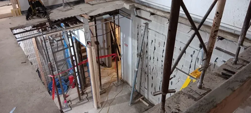A construction site showing a stairwell opening framed by scaffolding and safety barriers on a concrete floor.