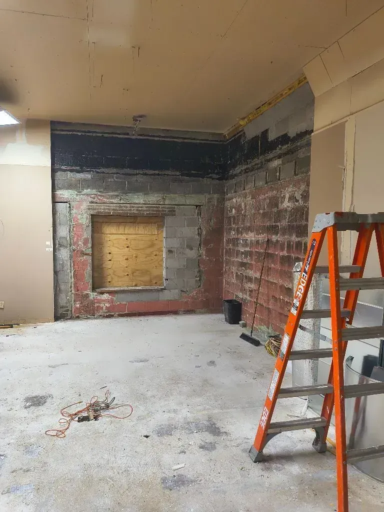 An interior room under renovation with exposed brick and block walls, a boarded-up window, and an orange stepladder.