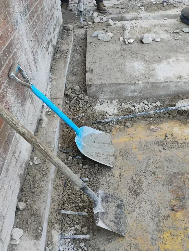 Two shovels rest against a brick wall at a dusty, active construction site with a concrete slab in the background.