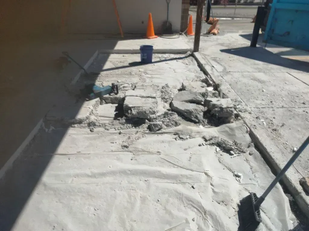A construction site showing a patch of broken concrete on a sidewalk, surrounded by safety cones and a blue bucket.