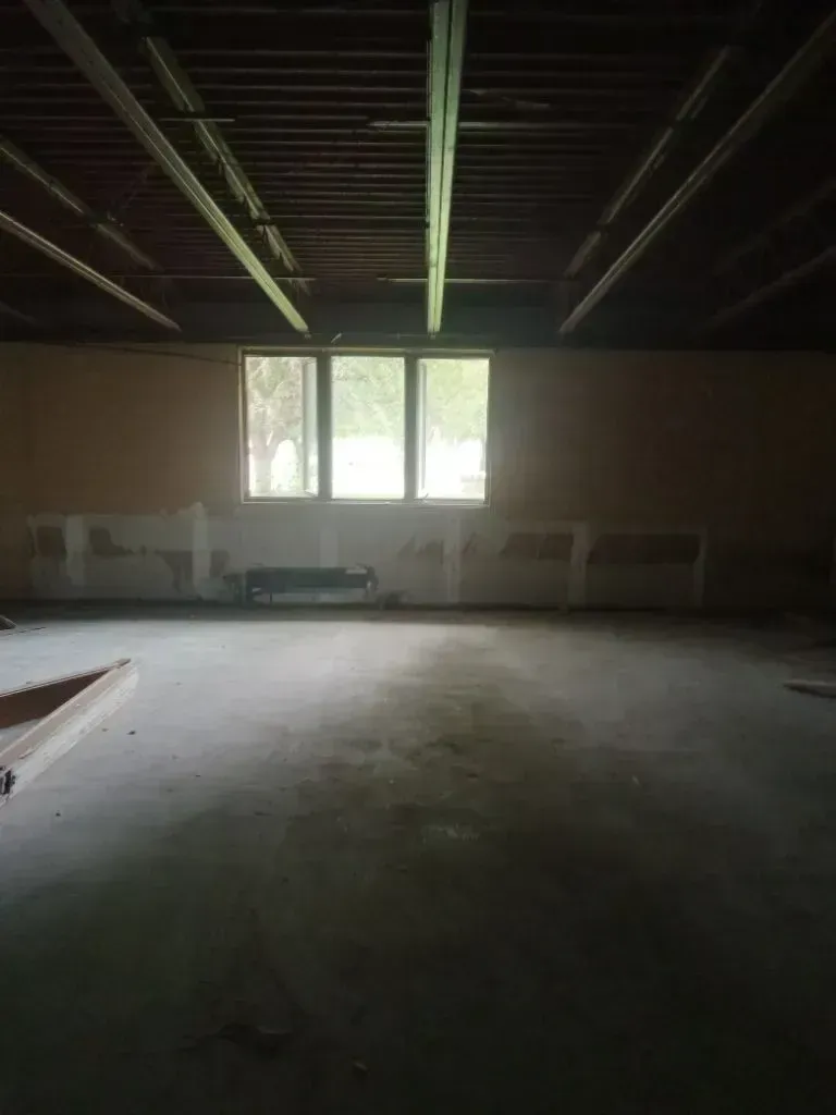 An unfinished room with exposed wooden ceiling beams, concrete floors, and a bright window centered on the back wall.