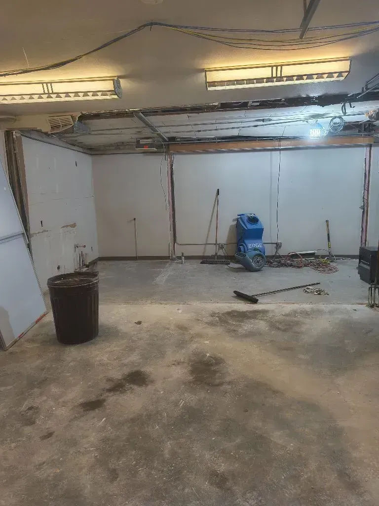 A dimly lit garage interior under renovation with bare white walls, a concrete floor, two ceiling lights, and tools.