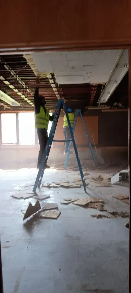 Two workers in neon vests stand on stepladders, removing ceiling panels in a room undergoing renovation.