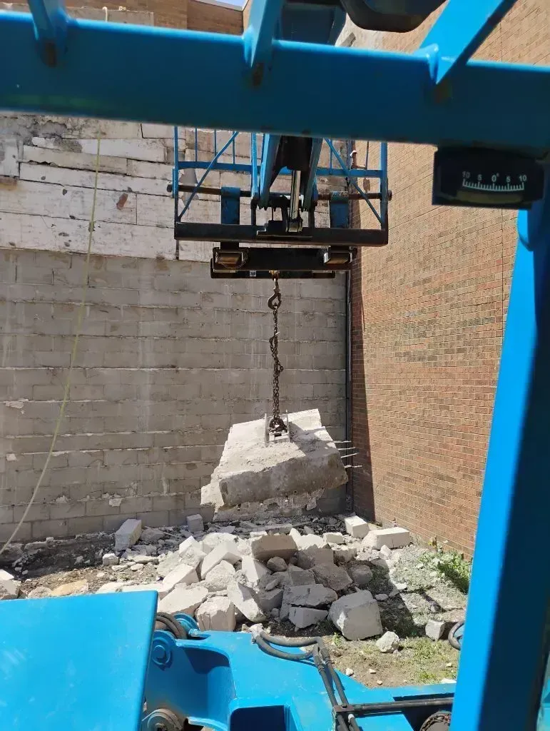 A blue construction crane lifting a large, heavy concrete block amidst debris at an outdoor demolition site.